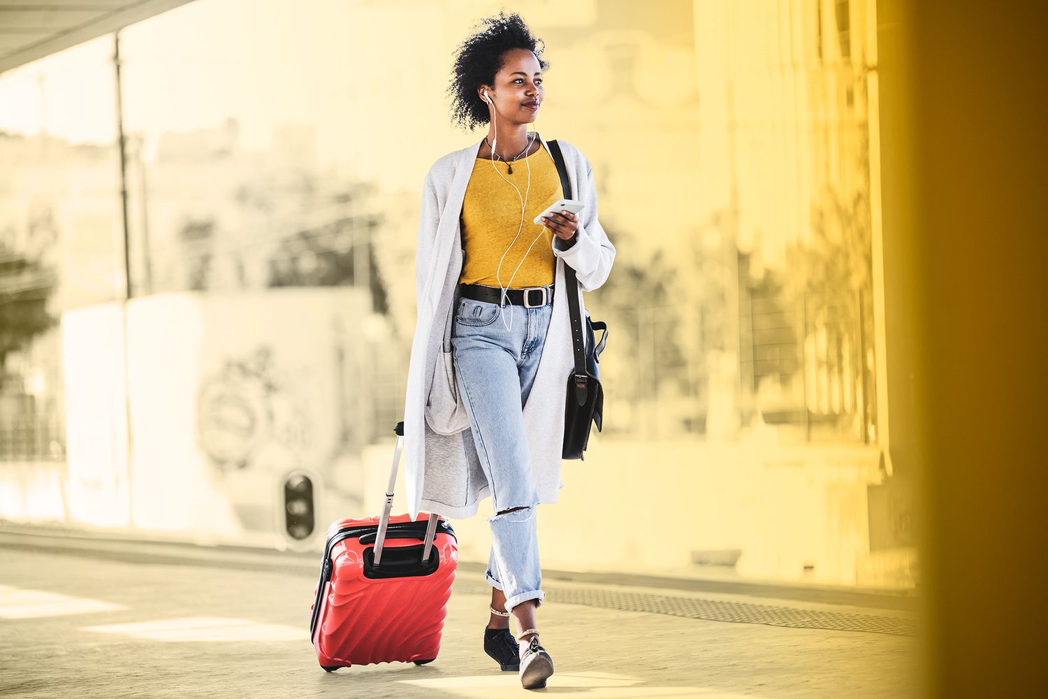 Una mujer joven con un teléfono móvil y auriculares caminando con una maleta