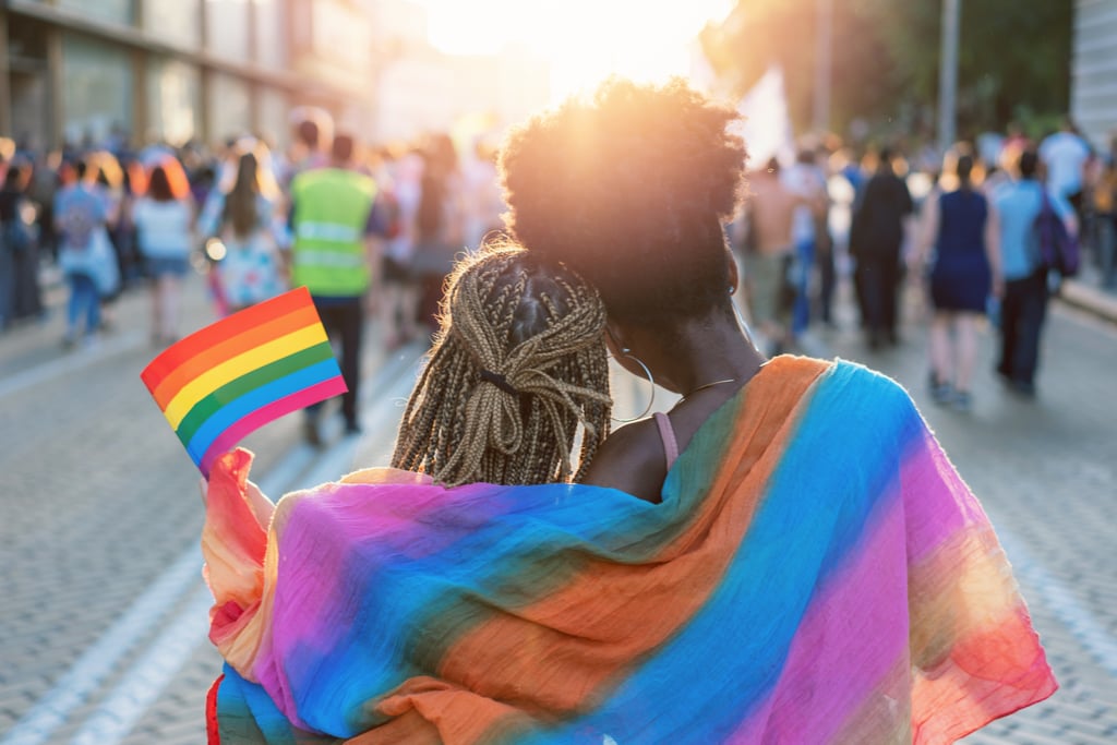 Vista trasera de una pareja joven caminando con un evento del Orgullo, abrazándose y ondeando una bandera del Orgullo.