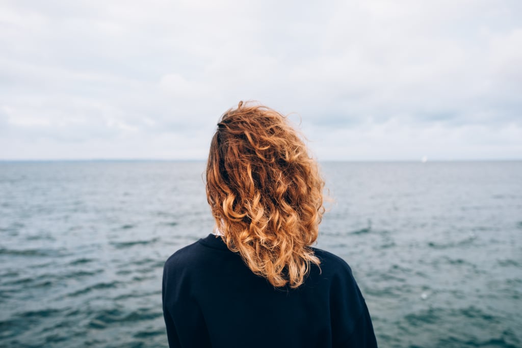 Una mujer con un cabello rizado rojo que está pensando en ello cerca del agua azul al aire libre. La parte trasera de una mujer joven mirando el mar.