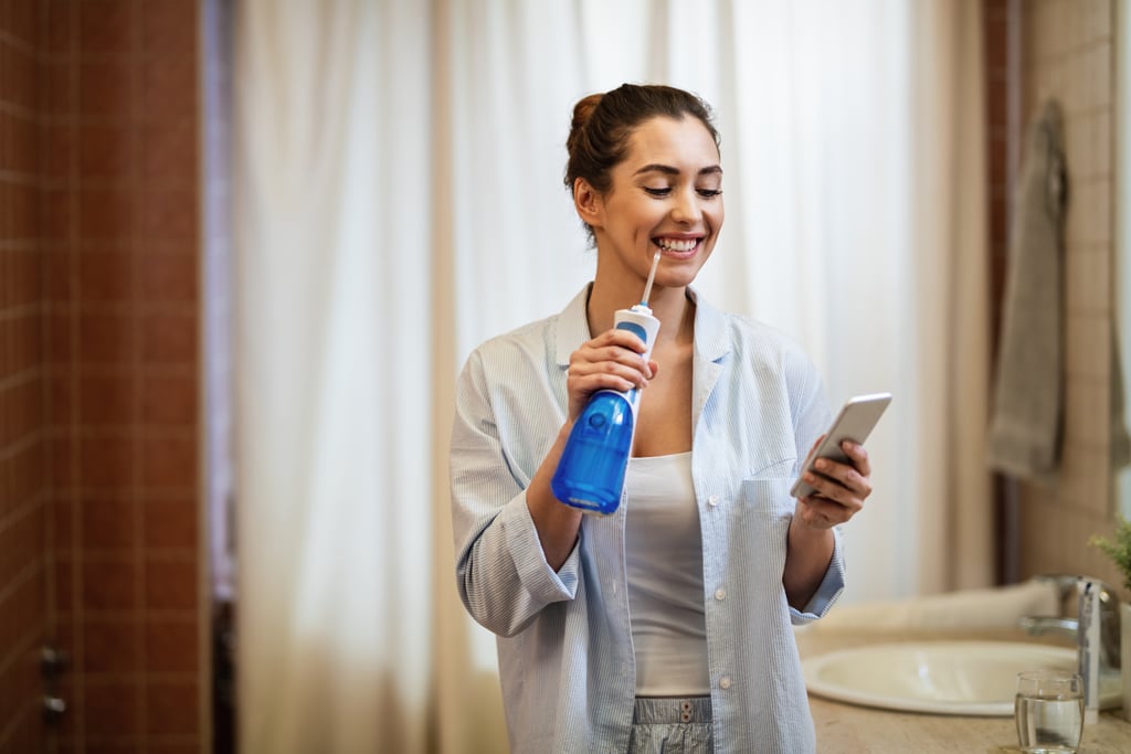 Una mujer joven y feliz que fluye agua con un hilo dental mientras envía un correo electrónico a una habitación móvil en el baño y se limpia los dientes.